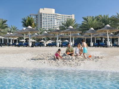 Kinder spielen am Sandstrand vor einem Hotel mit Sonnenliegen und Palmen.