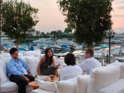 Vier Personen sitzen auf weißen Sofas in einem Freiluftbereich mit Blick auf einen Yachthafen bei Sonnenuntergang.