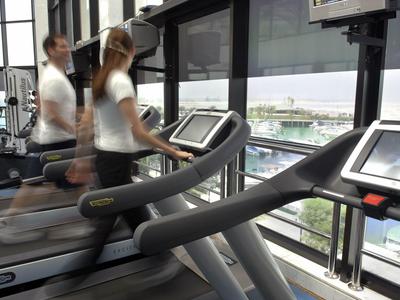 Zwei Personen laufen auf Laufbändern in einem hellen Fitnessraum mit großen Fenstern.