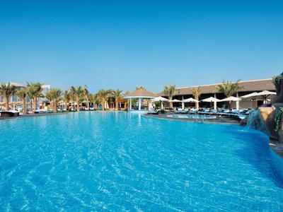 Großer Außenpool mit klarem blauem Wasser, umgeben von Liegestühlen und Palmen bei sonnigem Wetter.