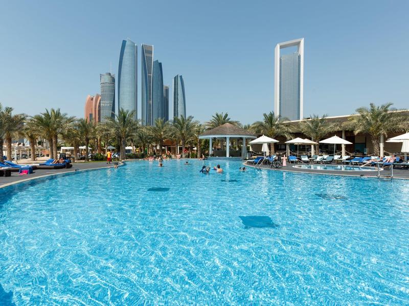 Großer Außenpool mit Sonnenliegen und Wolkenkratzern im Hintergrund bei sonnigem Himmel.