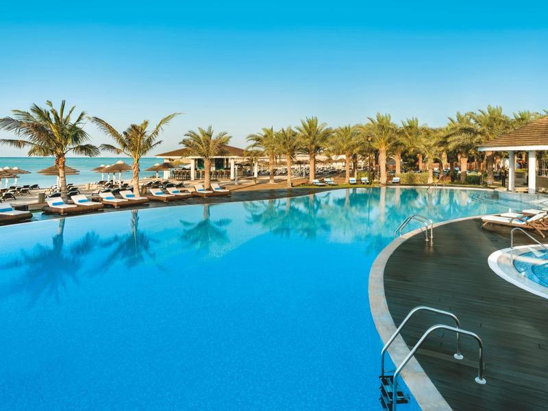 Großer Hotelpool mit Palmen und Liegestühlen neben dem Meer unter klarem Himmel.