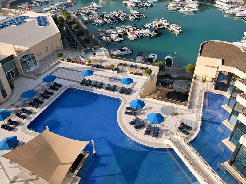 Blick auf einen Hotelpool mit blauen Liegestühlen und Blick auf einen Hafen mit Booten.