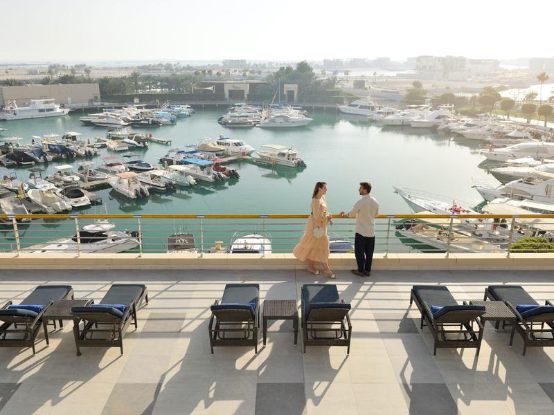 Paar auf Terrasse mit Liegestühlen mit Blick auf marina mit Booten bei Sonnenuntergang.