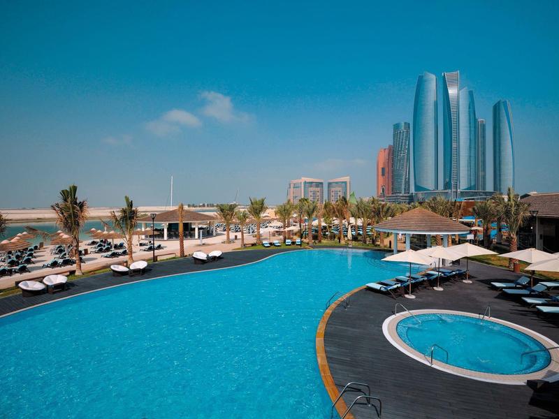 Großer Pool mit Liegestühlen und Hochhäuser im Hintergrund bei klarem Himmel.