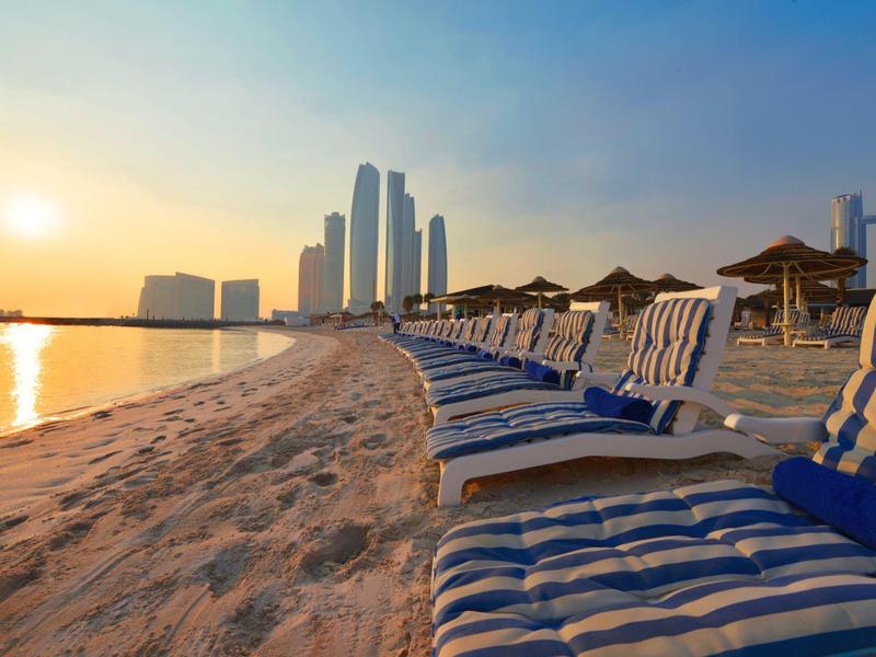 Sonnenuntergang am Strand mit Liegestühlen und moderner Skyline im Hintergrund.