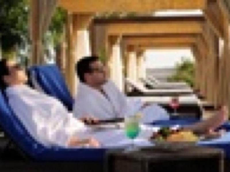 Zwei Personen entspannen in weißen Bademänteln auf blauen Liegen unter Holzpergolen.