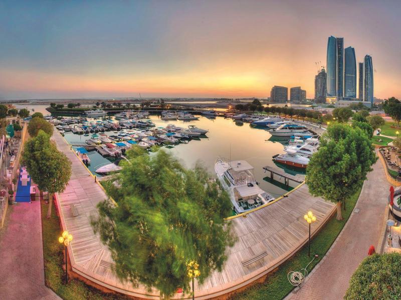 Panoramablick auf einen Jachthafen bei Sonnenuntergang mit hohen Gebäuden und einer Promenade.