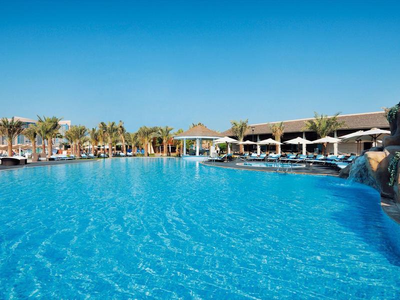 Großer Hotelpool mit klarem blauen Wasser, Palmen und Liegestühlen unter blauem Himmel.