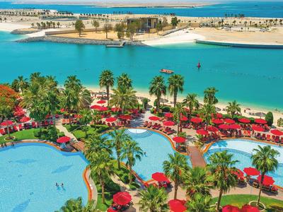 Blick auf tropischen Pool mit roten Sonnenschirmen und Palmen, dahinter türkisfarbenes Wasser und Sandstrand.