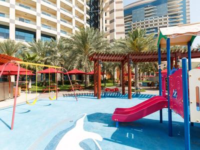 Bunter Kinderspielplatz mit Rutschen und Schaukeln vor einem Hotelgebäude und Palmen.