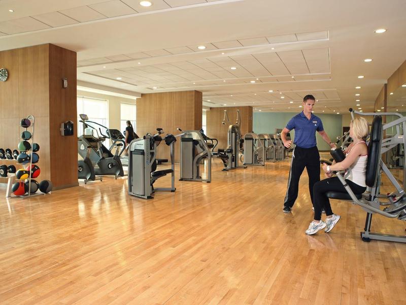 Hell beleuchteter Fitnessraum mit Holzboden, Gewichten, Trainingsgeräten und zwei Personen beim Training.