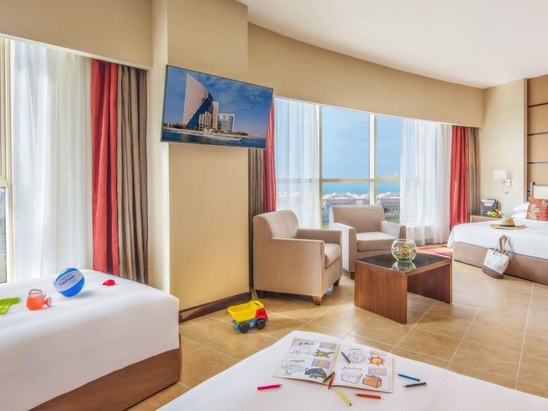 Helles Hotelzimmer mit großen Fenstern, zwei Betten, Sesseln, Tisch und Spielzeug auf dem Bett.