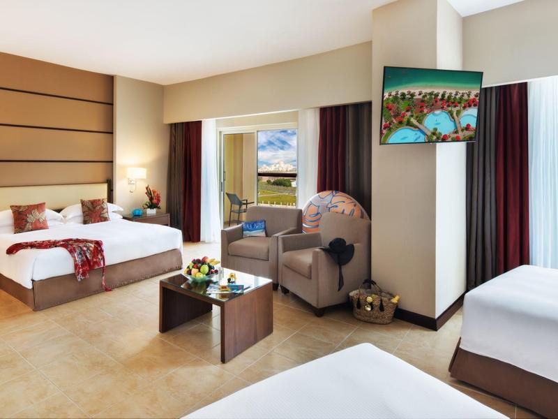 Helles Hotelzimmer mit zwei Betten, Sesseln, Tisch, Fernseher und großer Fensterfront.