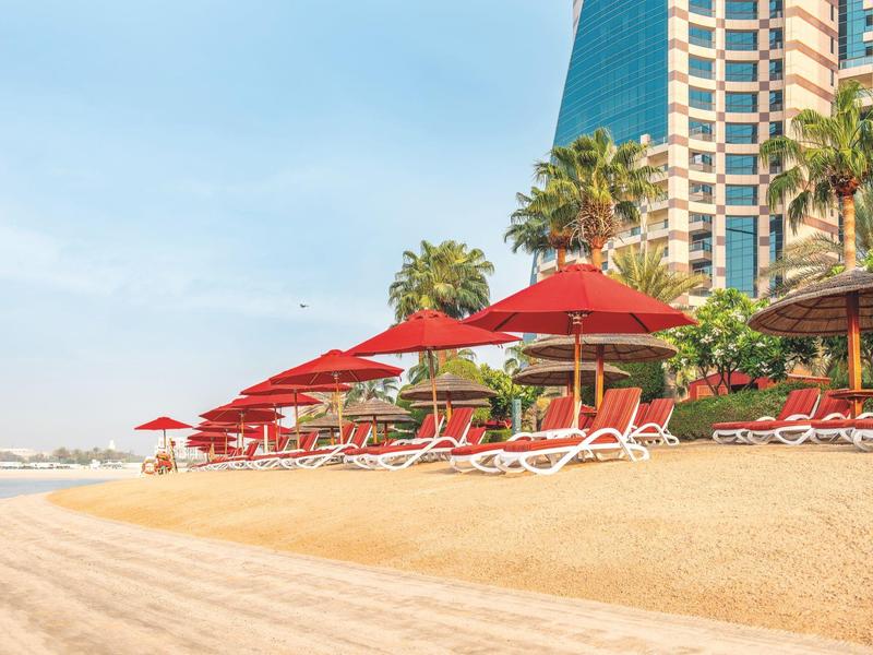 Sandstrand mit roten Sonnenschirmen, Liegestühlen und hohen Palmen vor Hochhauskulisse.
