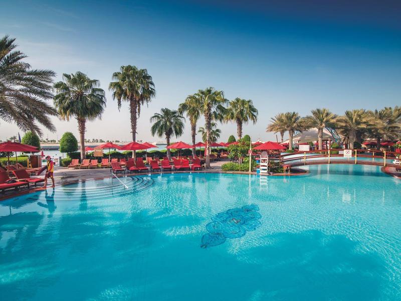 Großer Pool mit klar blauem Wasser, umgeben von Palmen und roten Sonnenschirmen bei Sonnenschein.