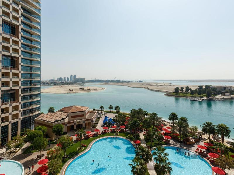 Blick auf Poollandschaft, Palmen und Wasserkanäle mit moderner Hochhausfassade links im Vordergrund.