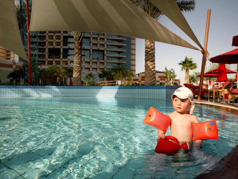 Ein Kind mit Schwimmflügeln in einem Außenpool, umgeben von Sonnenschirmen und Gebäuden.