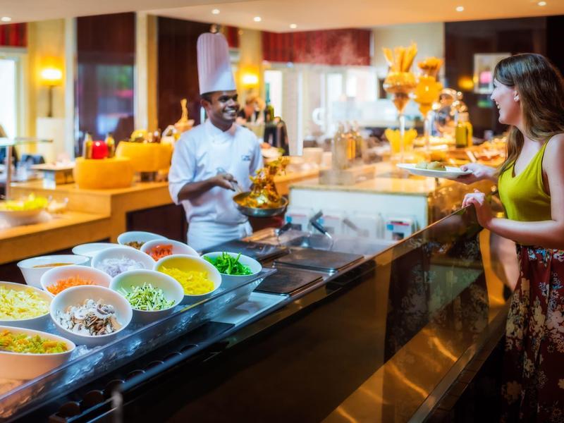 Buffet mit bunten Salaten und Gemüse, Koch in weißer Uniform bedient eine Frau in grünem Top.