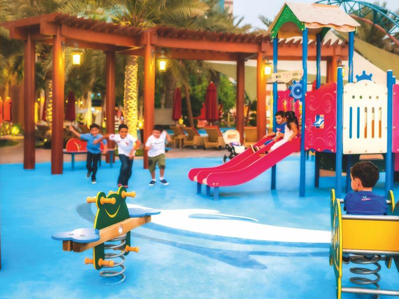 Bunter Spielplatz mit Rutsche, Schaukeln und Kindern auf blauem Boden unter Holzdach.