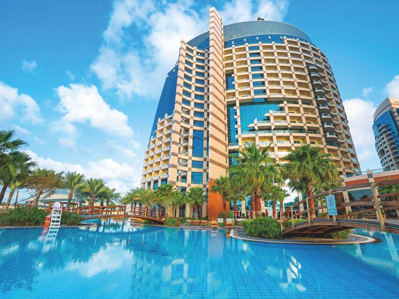 Moderner Hotelturm mit Glasfassade, Palmen und großem Swimmingpool unter blauem Himmel.