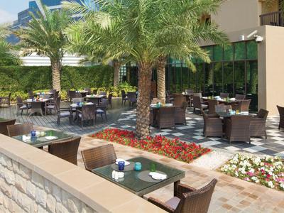 Außenbereich eines Cafés mit Tischen, Stühlen und Palmen, umgeben von Blumenbeeten.