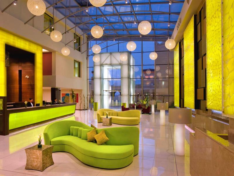 Moderne Hotellobby mit grüner, geschwungener Couch, gelben Wandpaneelen und leuchtenden Hängeleuchten.