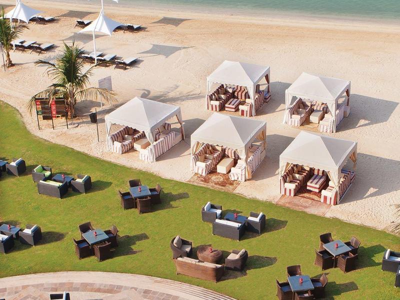 Ein Strand mit weißen Sonnenschirmen, Liegen und einem Grünbereich mit Sitzmöbeln in Reihen angeordnet.