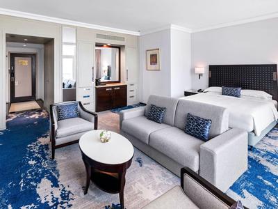 Geräumiges Hotelzimmer mit beigem Sofa, blau gemustertem Teppich und großem Bett.