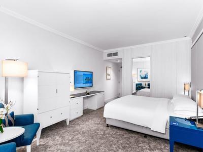 Helles Hotelzimmer mit großem Bett, blauem Sessel, Fernseher und modernem, weißen Mobiliar.