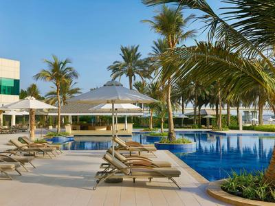 Moderner Außenpool mit Liegestühlen und Sonnenschirmen neben Palmen bei sonnigem Wetter.