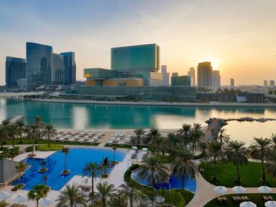 Blick auf einen Hotelpool mit Palmen und Stadtgebäuden im Hintergrund bei Sonnenuntergang.