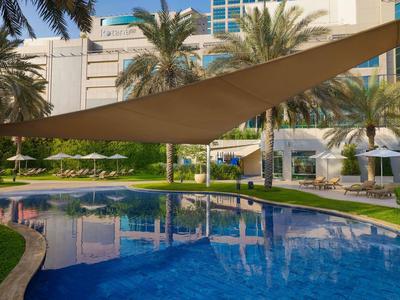 Moderner Hotelpool mit Sonnensegel, umgeben von Palmen und Liegestühlen.