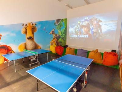 Innenraum mit zwei blauen Tischtennisplatten und Leinwand für Filme, umliegende bunte Cartoon-Wandbilder