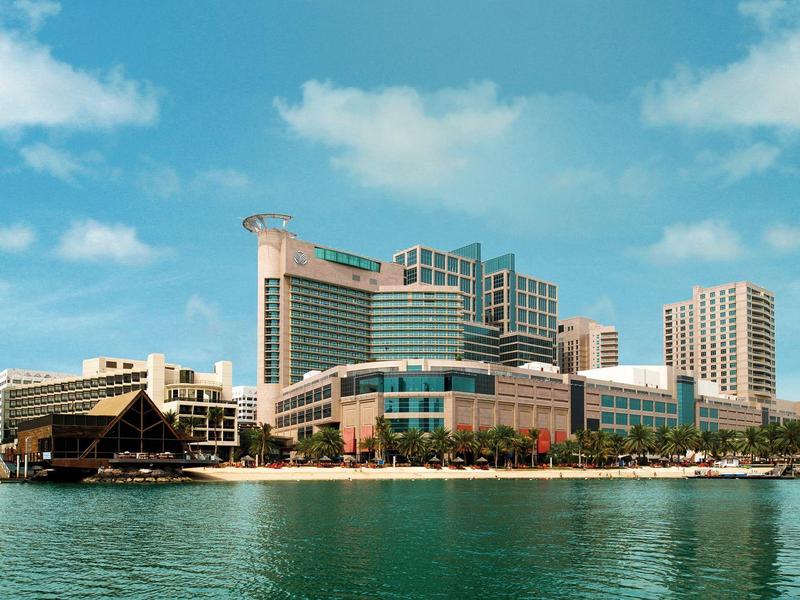 Hafenstadt mit modernen, hohen Gebäuden vor blauem Himmel und ruhigem Wasser im Vordergrund.