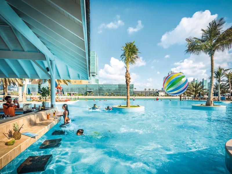 Großer blauer Pool mit Palmen und Sonnenschutz, Menschen schwimmen und spielen im Wasser.