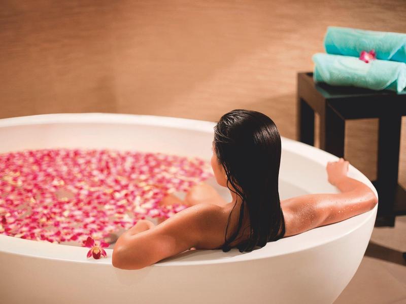 Frau entspannt in ovaler Badewanne mit Rosenblättern, neben grün gestapelten Handtüchern auf schwarzem Tisch.