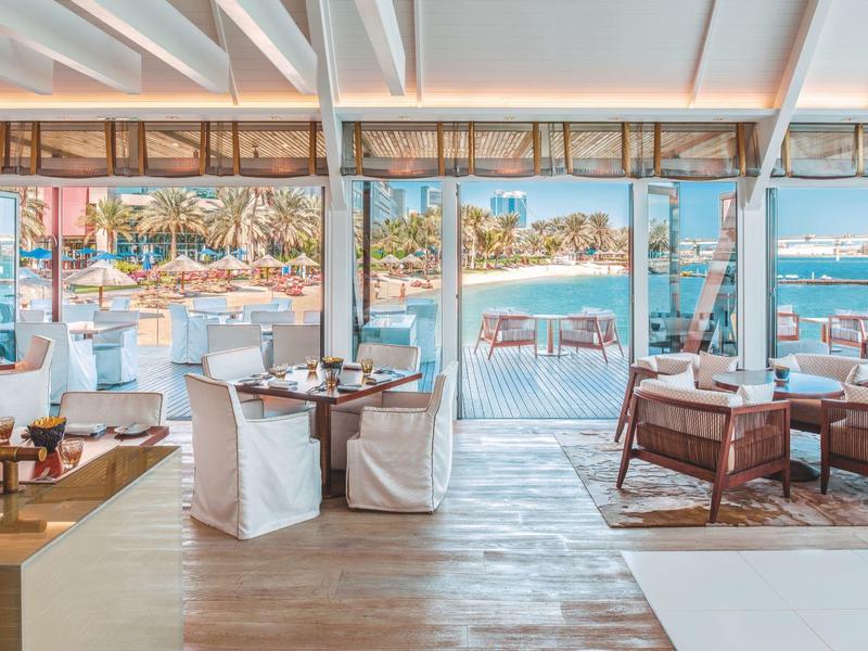 Helles Restaurant mit Holzfußboden, weißen Stühlen und Blick auf türkisfarbenes Wasser und Boote.