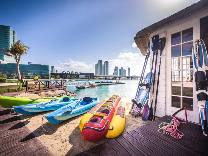 Kajaks in Blau, Gelb und Rot liegen auf Holzsteg, daneben Paddel vor Wasser und Wolkenkratzern bei Sonnenschein.