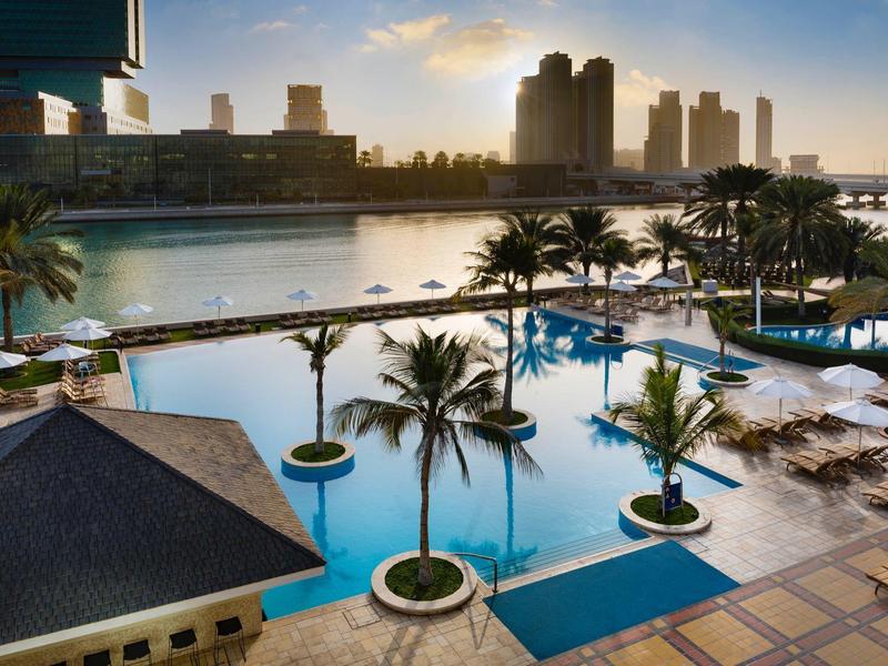 Großer Pool mit Palmen und Sonnenliegen, dahinter Fluss und moderne Hochhäuser bei Sonnenuntergang.