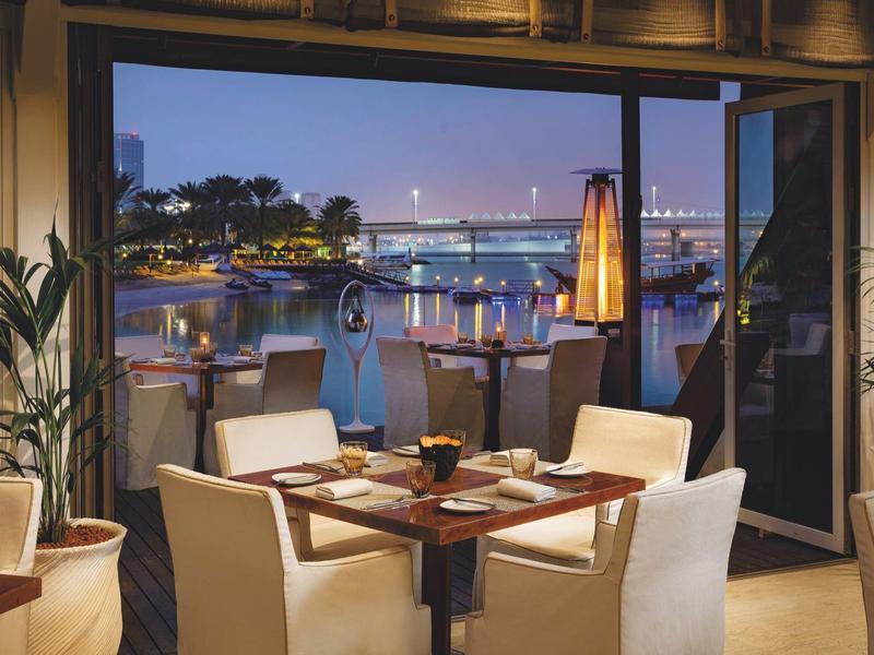 Gemütlicher Essbereich mit weißen Sesseln, Holztisch und Blick auf beleuchteten Hafen bei Dämmerung.