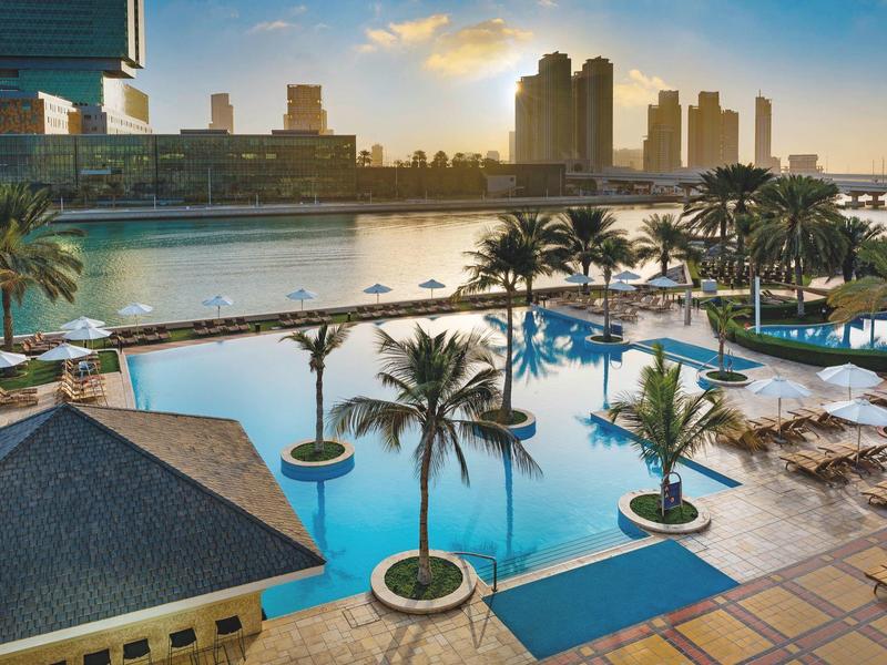 Ein großer Pool mit Palmen und Sonnenschirmen vor einer Stadt mit hohen Gebäuden bei Sonnenuntergang.