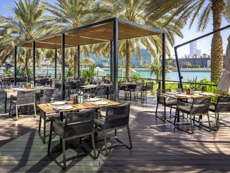 Offenes Restaurant mit Holzboden, Tischen, Stühlen unter Sonnendach und Palmen am Wasser.