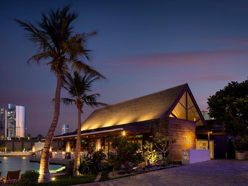 Ein beleuchtetes Holzhaus mit Palmen an einem ruhigen Wasserweg bei Dämmerung.