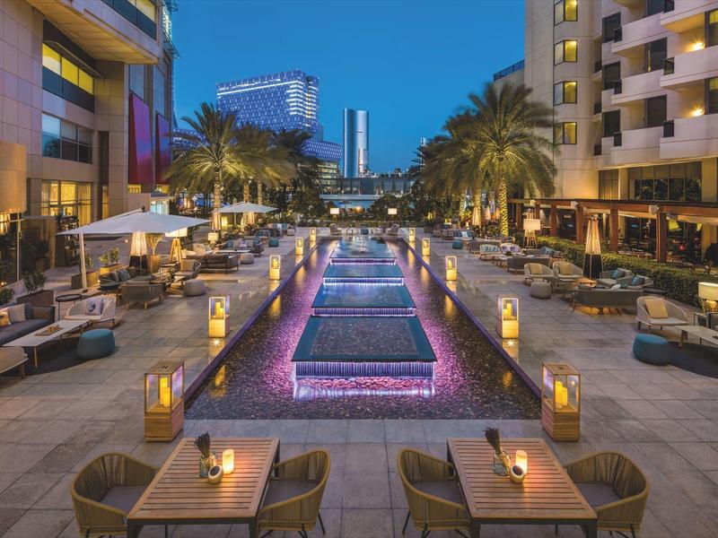 Moderner Außenbereich mit beleuchtetem Wasserbecken, Sitzgruppen und Palmen bei Dämmerung.