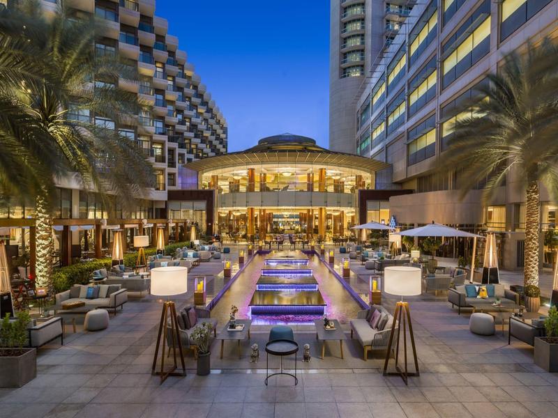 Moderner Außenbereich mit Pool, Palmen, Sitzgelegenheiten und beleuchtetem Restaurant bei Dämmerung.