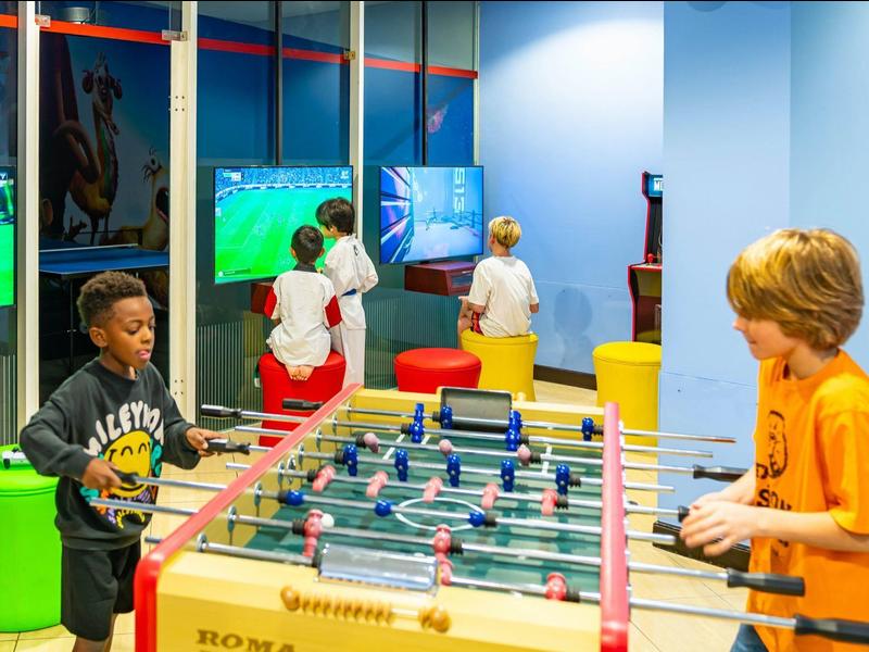 Kinder spielen in einem bunten Raum Tischfußball und Videospiele auf Bildschirmen an der Wand.