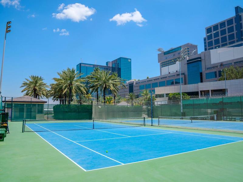 Freiluft-Tennisplatz mit blauem Belag, grüner Umgebung und Wolkenhimmel in urbaner Lage.