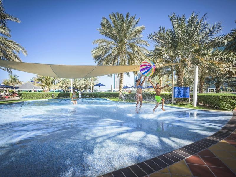 Kinder spielen im flachen Pool und werfen einen Ball unter Palmen bei blauem Himmel.