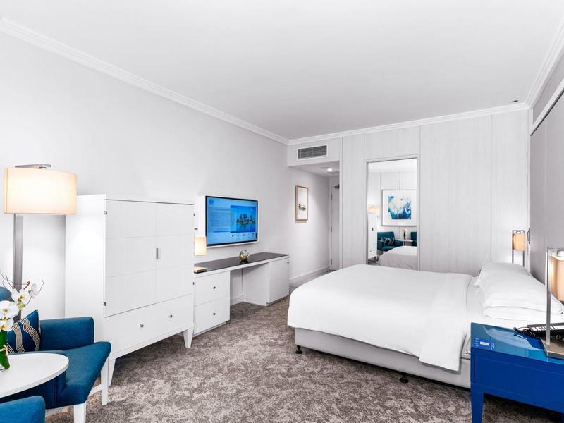 Helles Hotelzimmer mit weißem Bett, blauem Sessel und TV an der Wand.